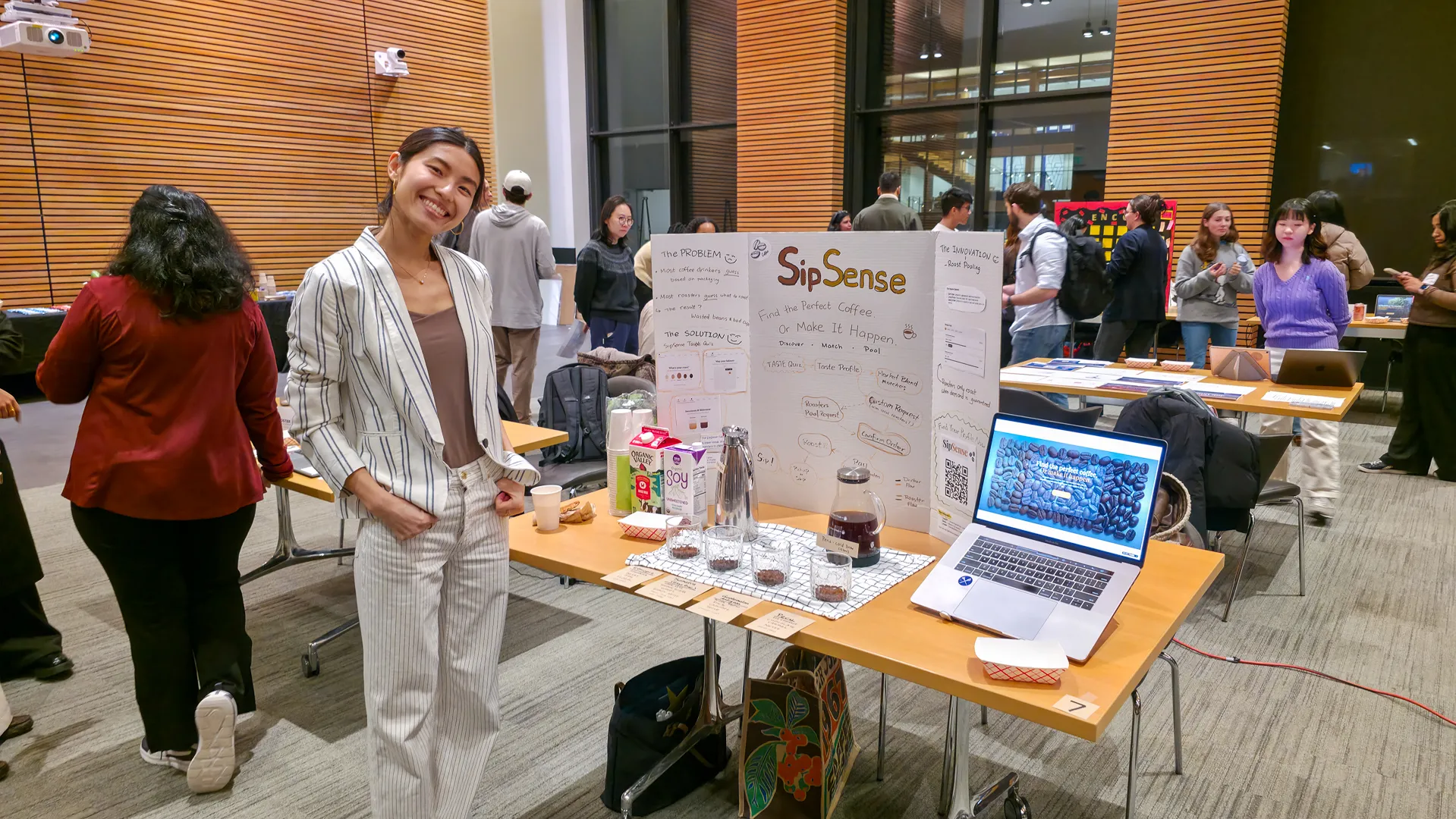 Student posing beside the SipSense demo table with project board, tasting setup, and laptop.